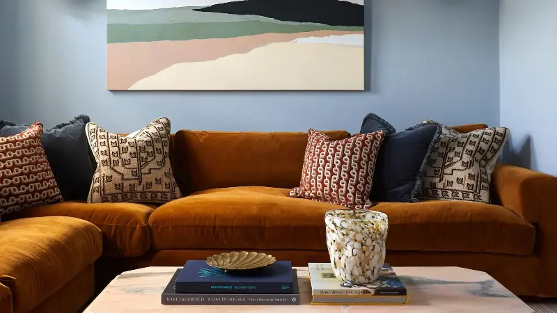 living room with blue walls, artwork, orange l-shaped sofa with lots of cushions and coffee table styled with books and vase