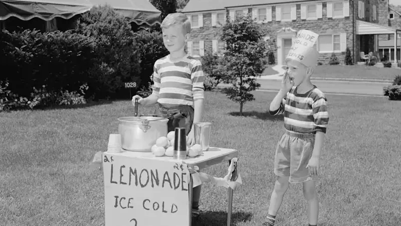 two young boys in striped shirts stand on a lawn selling lemonade for 2 cents