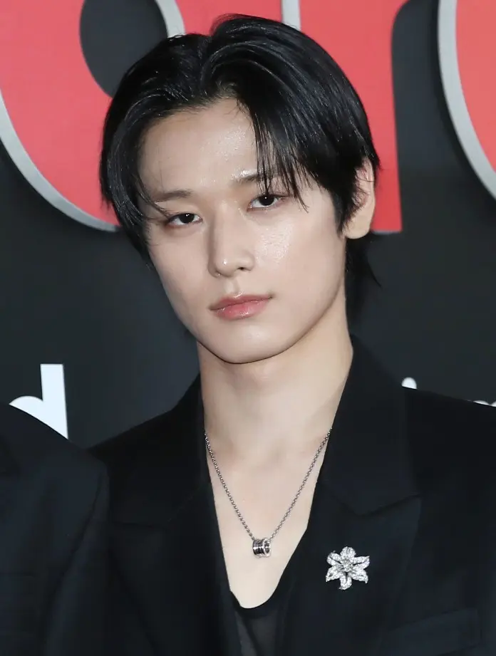 Close-up of Juyeon with dark, shoulder-length hair, wearing a black suit, looking directly at the camera. They have a silver necklace and a flower-shaped brooch on their lapel.