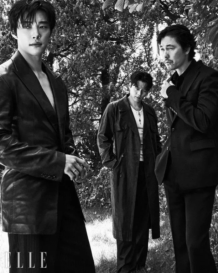A black and white image of Hyun Bin, Jung Woo-sung and Woo Do-hwan standing outdoors among trees. Woo Do-hwan on the left wears a dark, unbuttoned blazer and gazes directly at the camera. Hyun Bin in the center is dressed in a long coat and displays a contemplative expression. Jung Woo-sung on the right, dressed in a formal suit, appears thoughtful while adjusting his collar.