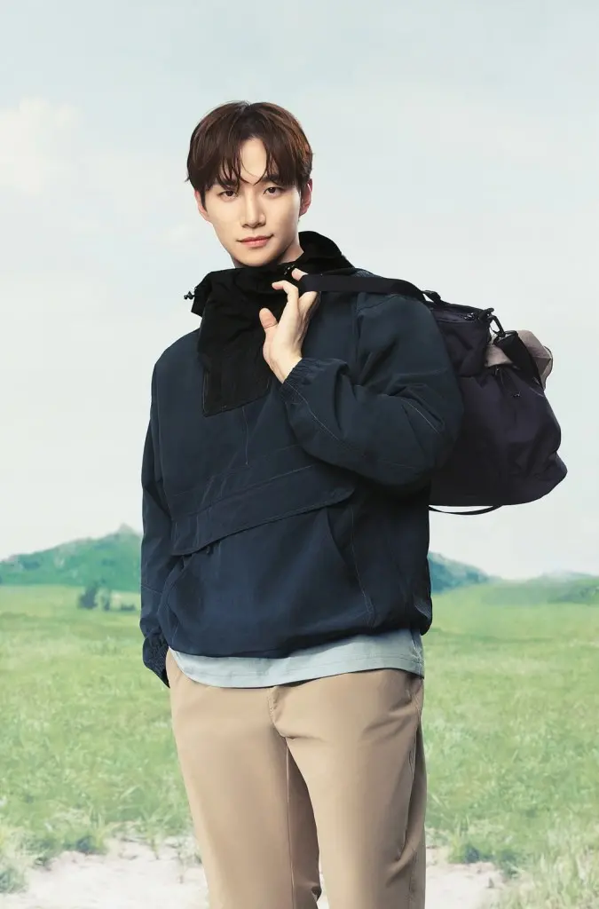 Lee Junho wearing a navy blue jacket and light beige pants stands outdoors, smiling while holding a backpack over one shoulder, with a grassy landscape and blue sky in the background.