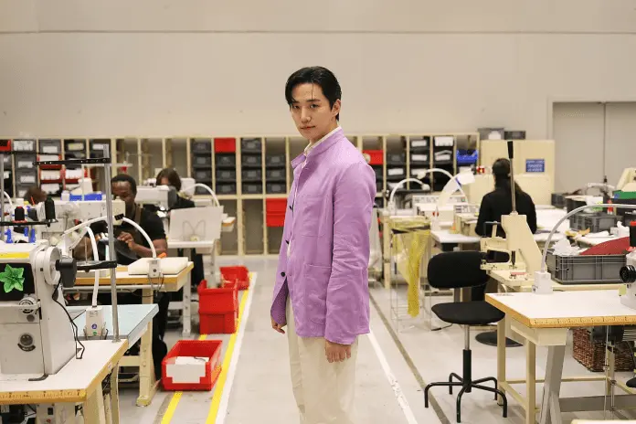 Lee Junho wearing a lavender jacket stands confidently in a sewing workshop, surrounded by sewing machines and workers engaged in garment production.