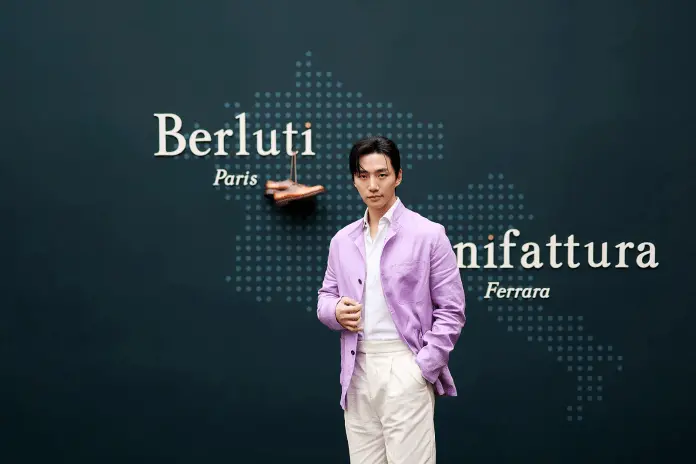 Lee Junho in a purple jacket stands confidently in front of a dark wall displaying the text 'Berluti Paris' and 'Manifattura Ferrara'.