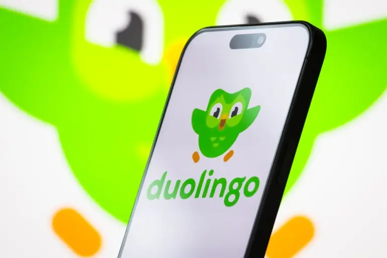 CHONGQING, CHINA - APRIL 26: In this photo illustration, the logo of Duolingo, Inc. is displayed on a smartphone screen, with the company's green owl mascot, Duo, visible in the background, on April 26, 2025, in Chongqing, China. Duolingo is an American edtech company that provides a popular language learning platform through its mobile app, known for gamified lessons and AI-powered instruction. (Photo illustration by Cheng Xin/Getty Images)