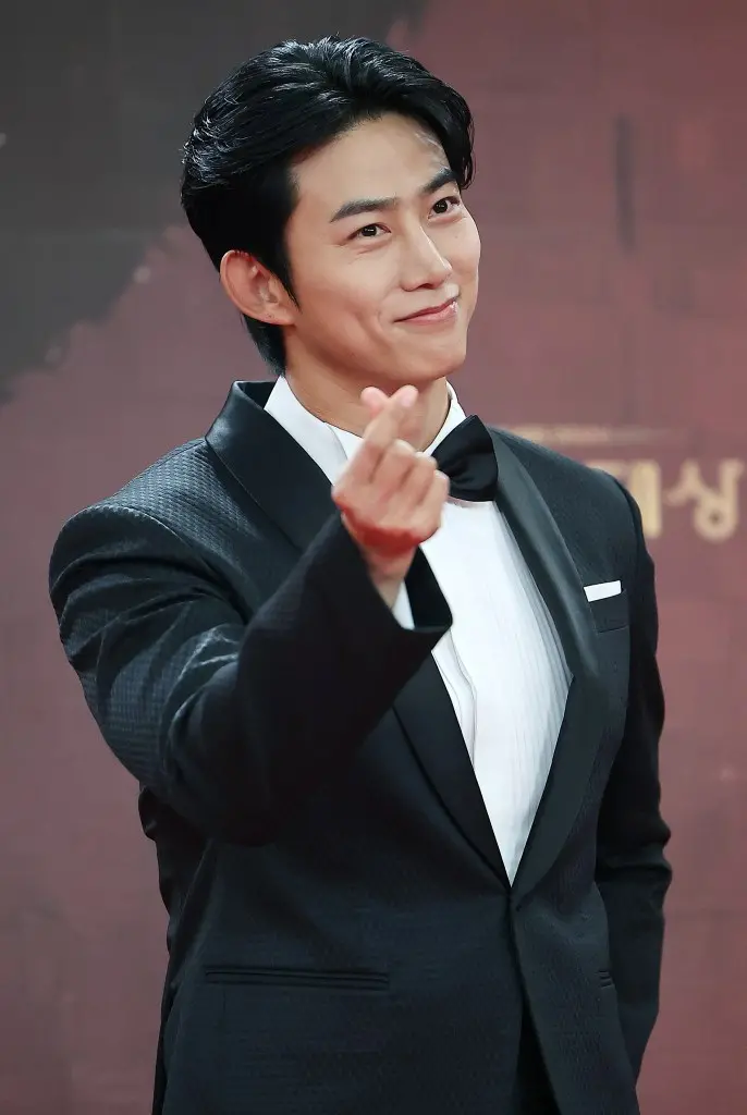 Ok Taecyeon in a tuxedo smiling and making a finger heart gesture, posing at an event.