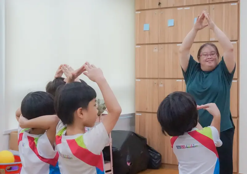 A trainer demonstrates a hand gesture activity with a group of preschoolers during a lesson