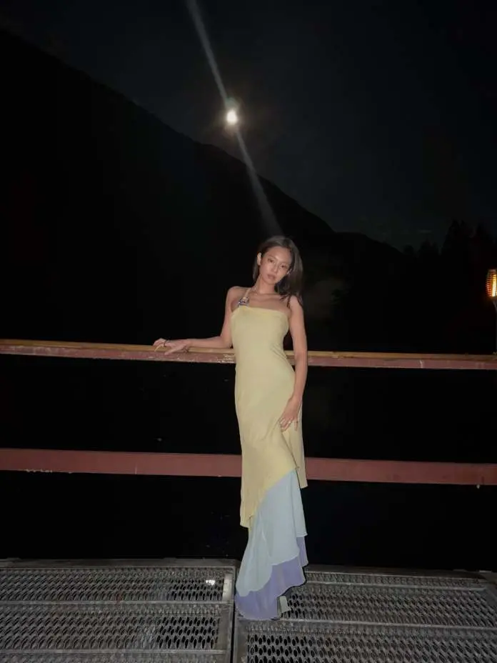 Jennie stands on a metal platform at night, wearing a long dress that is yellow at the top and transitions to soft pastel colors at the bottom. The moon is visible in the background, illuminating the scene.