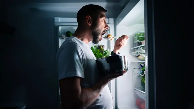 Hungry man eating food at night from open fridge. Man taking midnight snack from refrigerator;:
