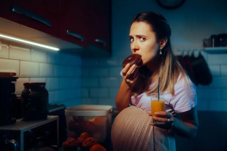 Pregnant Woman Eating Chocolate Doughnuts Drinking Orange Juice. Funny hungry mother to be snacking during nighttime