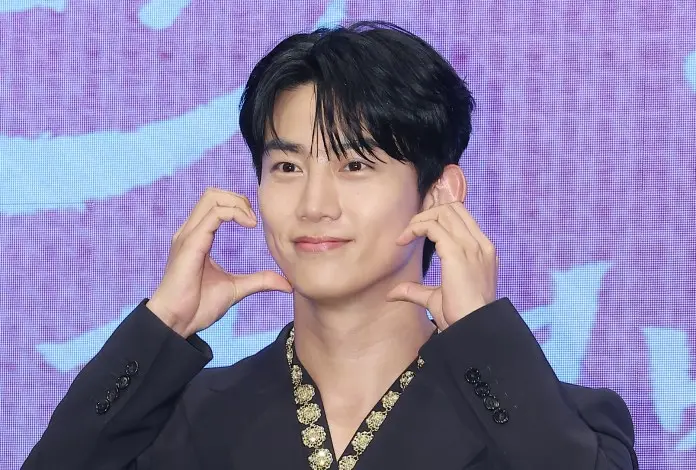 Ok Taecyeon is making a cheerful expression, holding their hands to their face in a playful gesture, wearing a black outfit with a decorative trim.