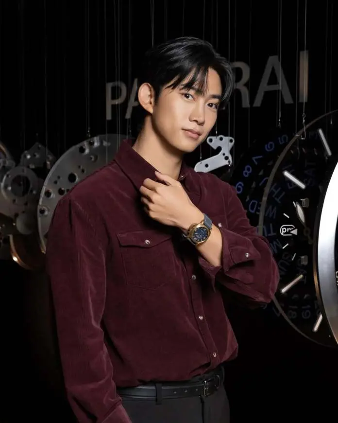 Ok Taecyeon wearing a burgundy corduroy shirt poses confidently, adjusting his collar while showcasing a stylish watch. The background features watch components and gears.