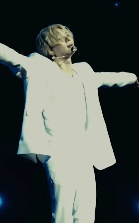 A performer in a white suit with arms outstretched, singing on stage under bright lighting.