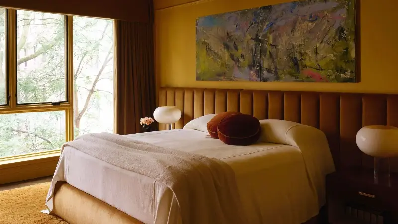 A modern bedroom with ochre color-drenched walls with decorative moldings, a large bed with neutral bedding, and a caramel-brown elongated headboard. A warm brown textured rug and large windows.