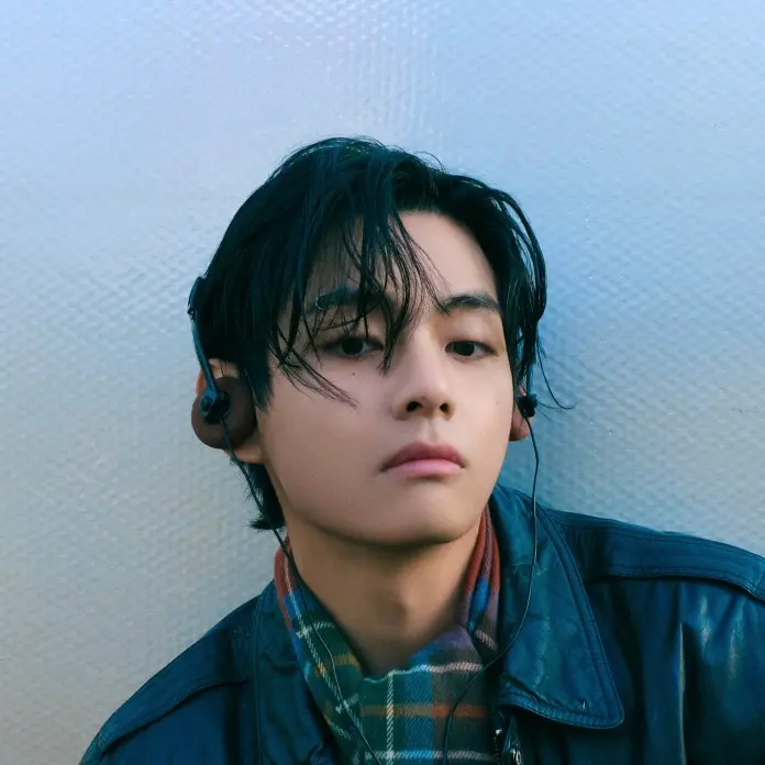 BTS V with dark hair and headphones, wearing a leather jacket and a patterned scarf, gazes thoughtfully at the camera against a textured background.