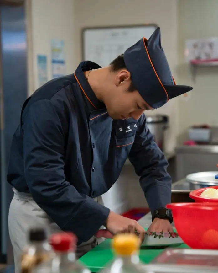 Park Ji-hoon wearing a dark blue uniform and hat is focused on chopping vegetables on a green cutting board in a kitchen setting.