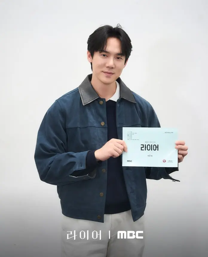 Yoo Yeon-seok dressed in a blue jacket and sweater is smiling while holding a script for a project titled '라이어' against a plain white background.