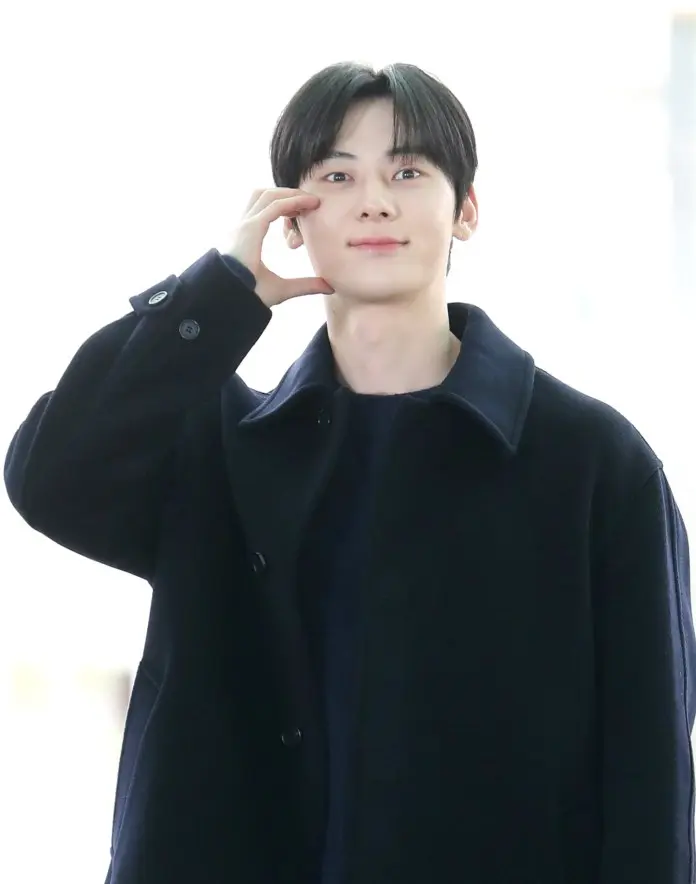 Hwang Min-hyun smiling and making a playful gesture with his hand while wearing a dark coat.