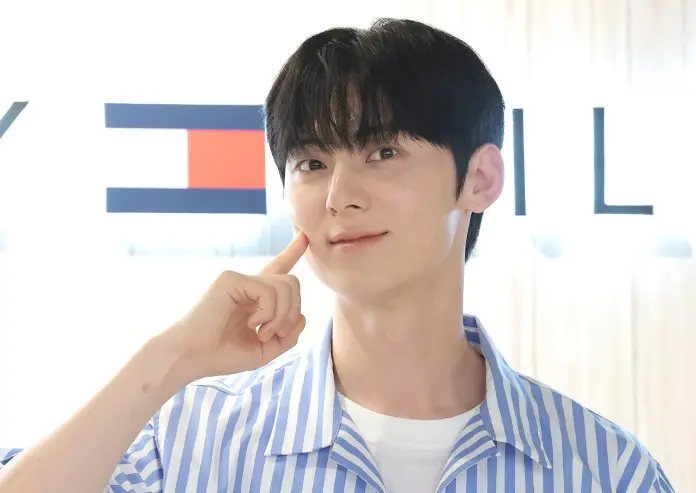Hwang Min-hyun with black hair, wearing a striped blue and white shirt, smiling and resting a finger on their cheek against a softly blurred background.