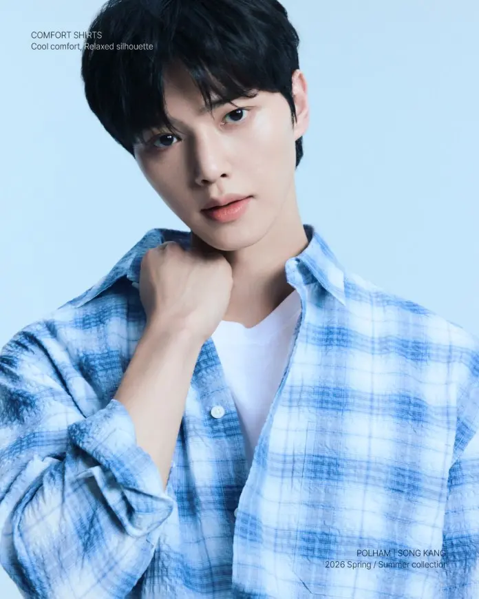 Song Kang  posing confidently in a blue plaid shirt over a white t-shirt, with a soft blue background.
