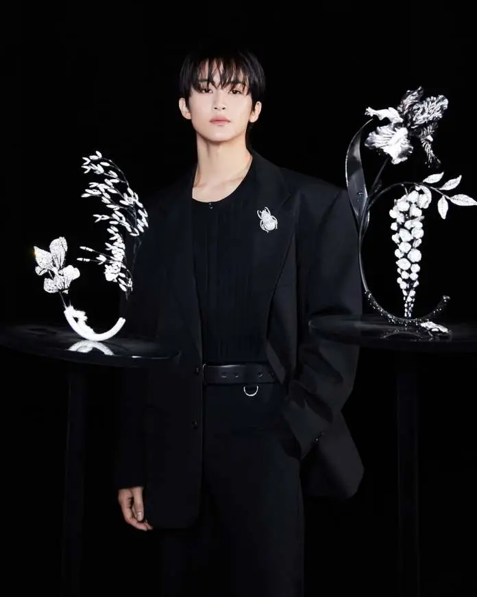 Mark Lee dressed in a black suit stands confidently against a dark background, with two decorative displays featuring floral designs on either side.