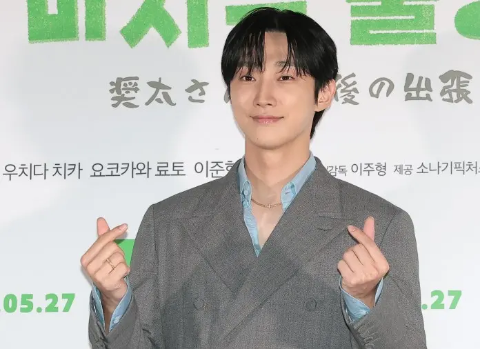 Jinyoung with dark hair wearing a gray suit and light blue shirt makes a heart gesture with their hands, smiling in front of a promotional backdrop.