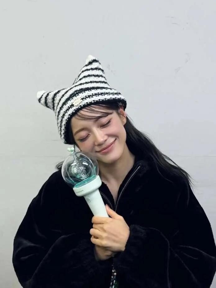 Kim Sejeong  smiling wearing a striped cat-ear hat and a black jacket, holding a lightstick close to their face against a simple gray background.