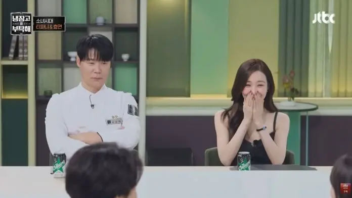 A man in a chef's uniform sits with arms crossed, looking pensive, while a woman in a black dress sits beside him, smiling and covering her mouth with her hands in surprise or laughter.