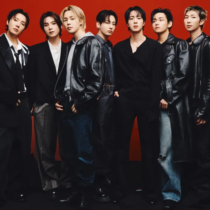 A group of seven individuals posing together against a red background, dressed in stylish, modern outfits, showcasing a mix of black leather, blazers, and denim. Their expressions are confident and serious.