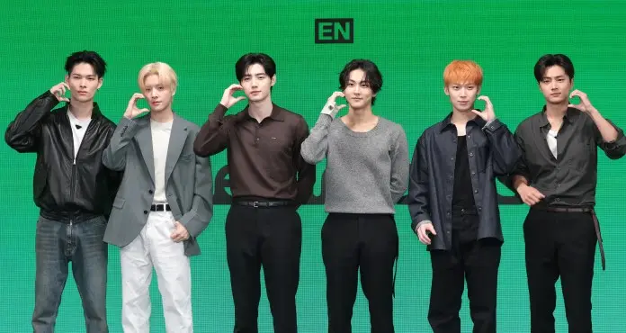 ENHYPEN  members posing during a promotional event, each making a hand gesture by placing their hand to their ear, with a green background.
