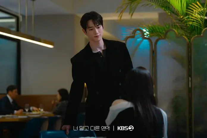 Seo Kang-joon in a stylish black jacket leans slightly over a table, looking at a woman seated in a restaurant. Soft lighting and indoor plants create a cozy atmosphere.