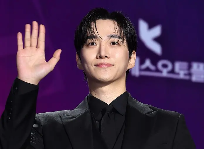 Lee Junho in a black suit smiling and waving, with a purple background.