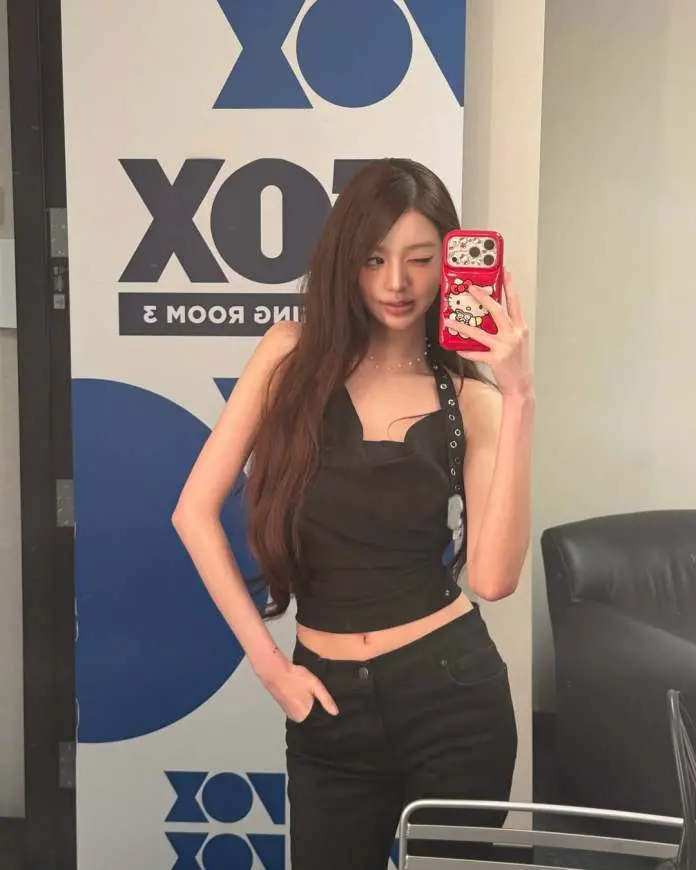 Jang Wonyoung taking a selfie in front of a mirror, wearing a black sleeveless top and black pants, with long hair and a wink. The background features a blue and white logo design.