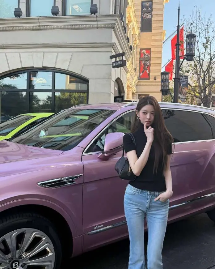 Jang Wonyoung in a black top and light blue jeans stands next to a pink luxury SUV, posing thoughtfully with her finger near her chin. The background features a stylish building and banners, indicating a lively urban setting.