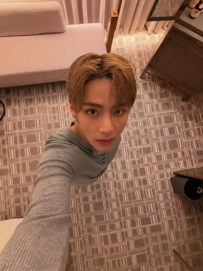 A person taking a selfie in a hotel room, wearing a gray long-sleeve shirt, with light brown hair and a neutral expression. The background features a patterned floor, a sofa, and a side table.
