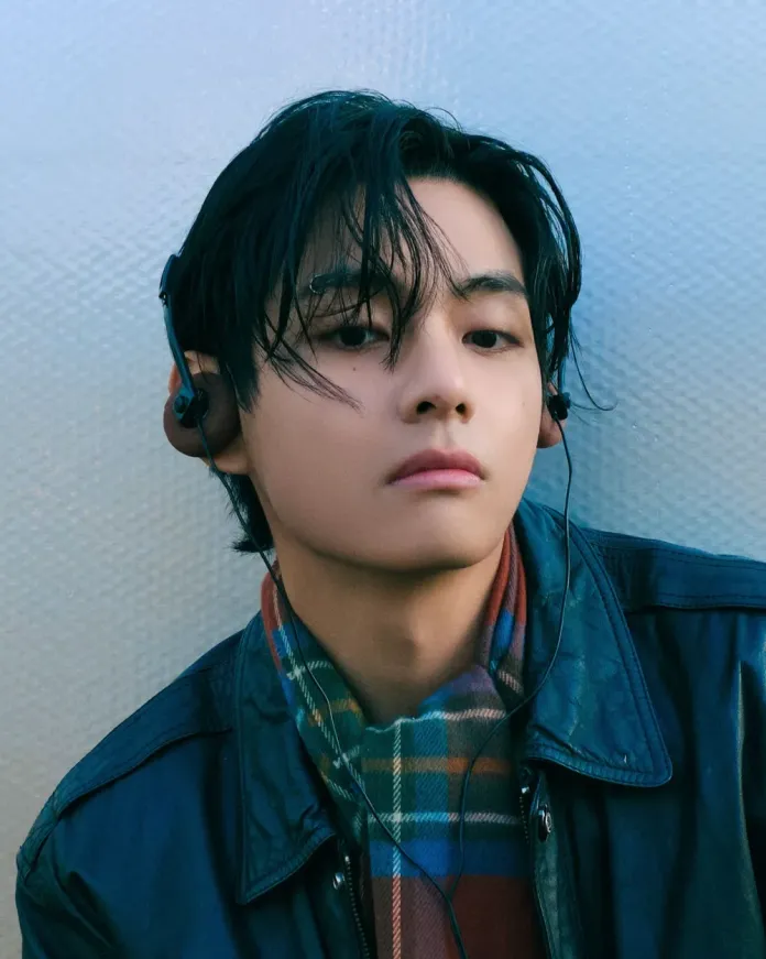 V with dark hair wearing headphones and a leather jacket, looking thoughtfully at the camera, with a checkered scarf around his neck.
