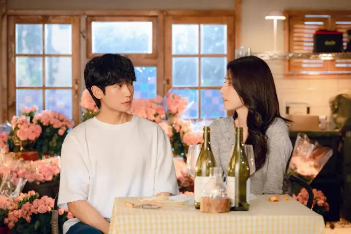 A young man and woman sitting at a table, engaged in conversation, surrounded by blooming flowers and wine bottles in a cozy interior setting.