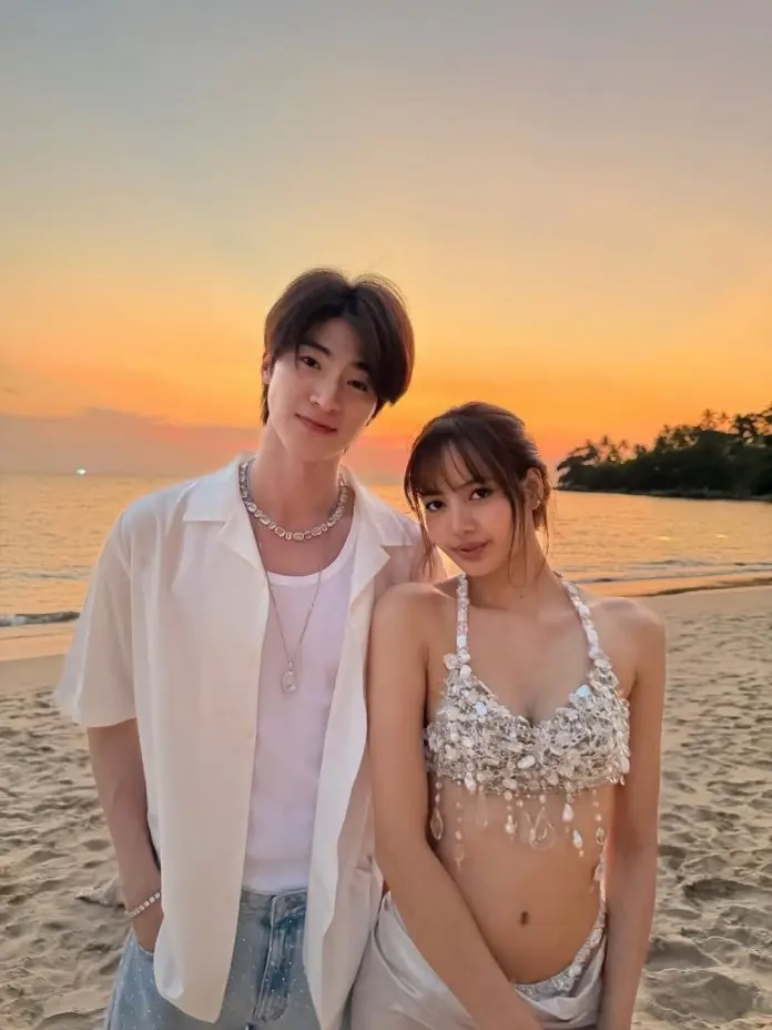 Lisa and Thai Actor Blue Pongtiwat poses for a photo on the beach at sunset, with a colorful sky in the background. The man is wearing a white shirt and denim jeans, while the woman is dressed in a sparkling, intricately designed top and a revealing outfit.
