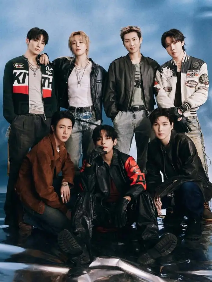 A group photo featuring BTS members posing together. They are dressed in a variety of stylish outfits, including jackets and casual wear, with a backdrop of soft blue clouds. The composition highlights their diverse fashion choices and expressions.