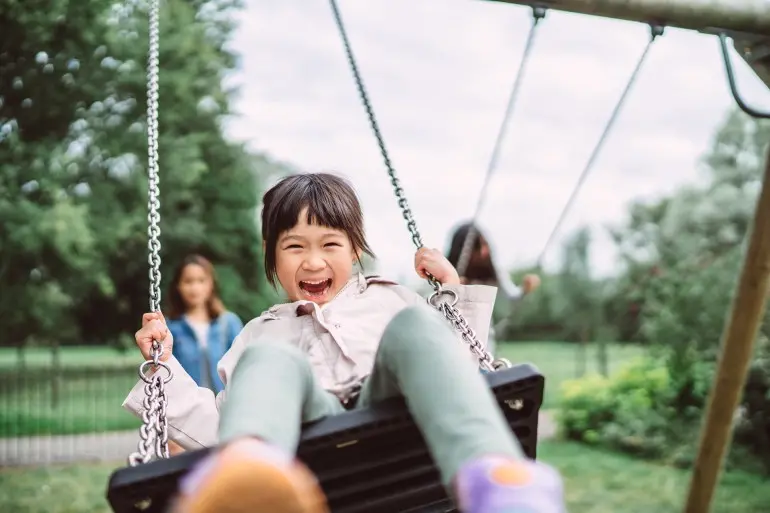 Lovely little girl smiling at the camera while playing on a swing set in playground joyfully.