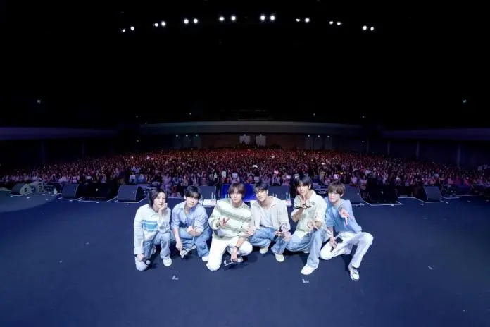 NCT DREAM members pose in front of a large audience at a concert, with a sea of fans in the background holding signs and lights.