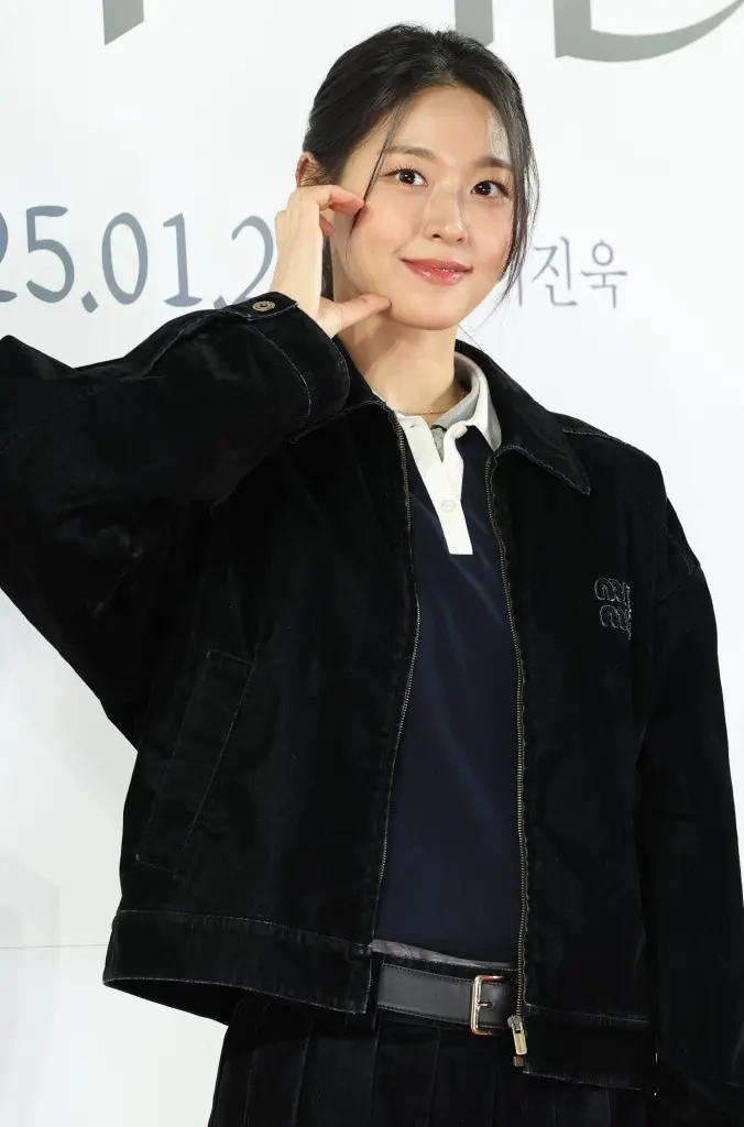 Seolhyun with long hair styled casually is posing and smiling while wearing a black corduroy jacket over a polo shirt. She is making an expressive hand gesture near her face against a simple white background.