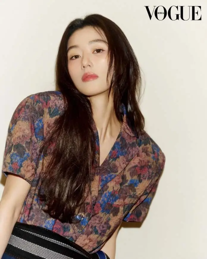Jun Ji-hyun with long dark hair poses stylishly, wearing a patterned shirt and a wide belt, against a light background. The image is featured in Vogue Korea.