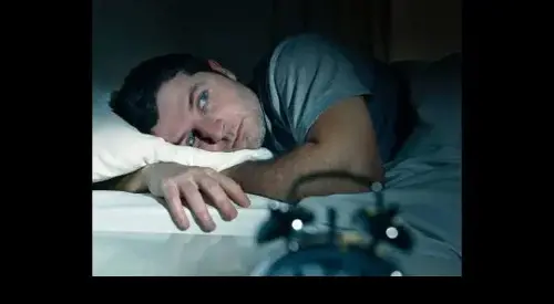 Hombre acostado en la cama con insomnio, mirando fijamente al vacío mientras un reloj despertador marca la madrugada, reflejando los efectos del trabajo nocturno en el sueño.
