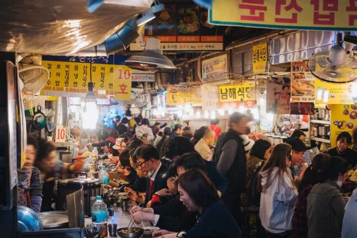 A bustling food market scene filled with people dining at various tables, bright yellow signs displaying menu items in the background.