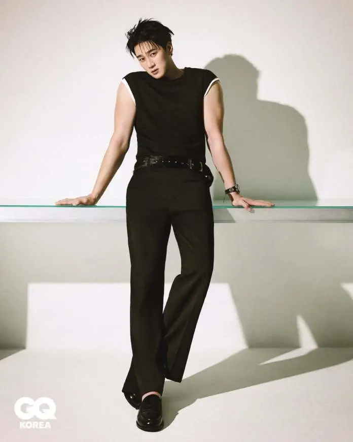 Ahn Bo-hyun posing confidently while leaning on a transparent glass surface, wearing a fitted black sleeveless shirt and black trousers. The background is minimalistic with soft lighting casting shadows.