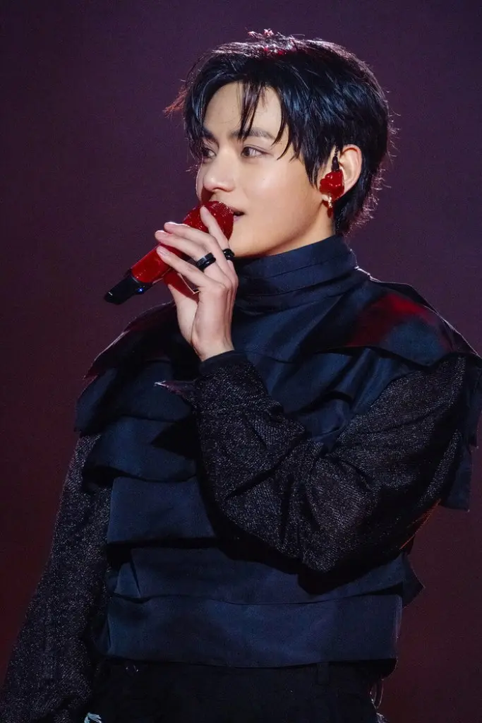 V on stage, wearing a black outfit with ruffled details, holding a red microphone and showcasing a stylish hairstyle against a blurred background.