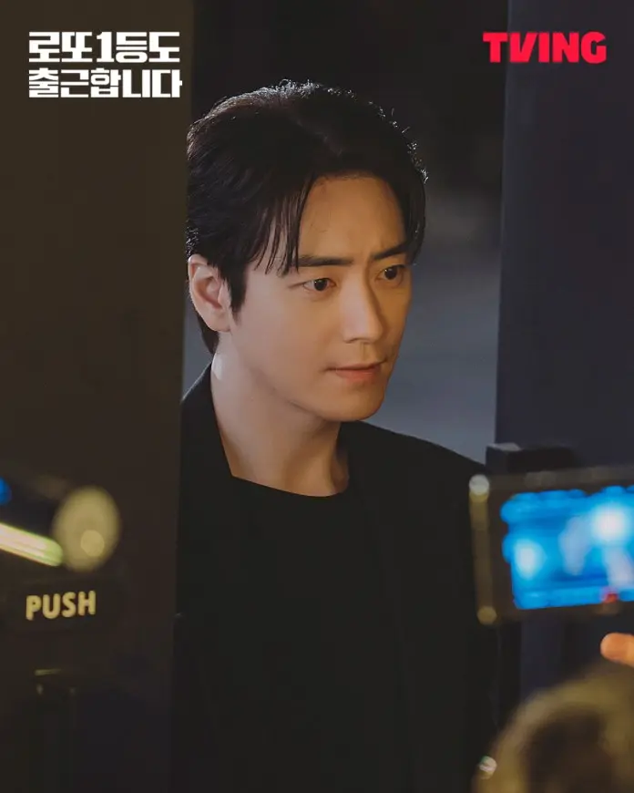 A close-up shot of Lee Joon-hyuk with dark hair, wearing a black shirt and jacket, looking intently with a serious expression. A camera is visible in the foreground capturing the moment.