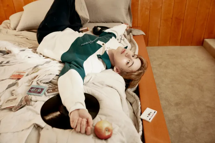 BTS V lying on a bed with a casual outfit, surrounded by cassette tapes, a vinyl record, and an apple, set against a wooden wall.