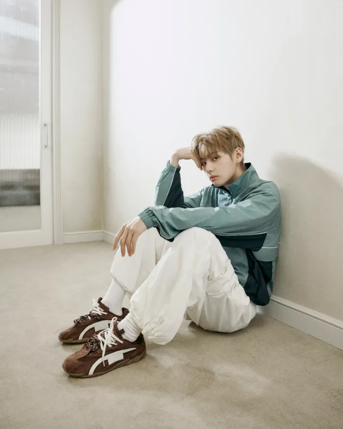 BTS V in a teal jacket and white pants sitting on the floor, looking contemplatively towards the camera, with brown sneakers on his feet and a minimalist background.