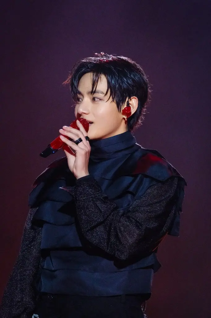 A vocalist holding a red microphone while performing on stage, wearing a black outfit with ruffled details and red accents in their hair.